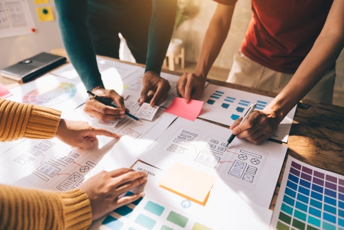 An overhead view of a collaborative design team working around a wooden table, reviewing paper wireframes, color swatch palettes, and sticky notes for a UX web design project.